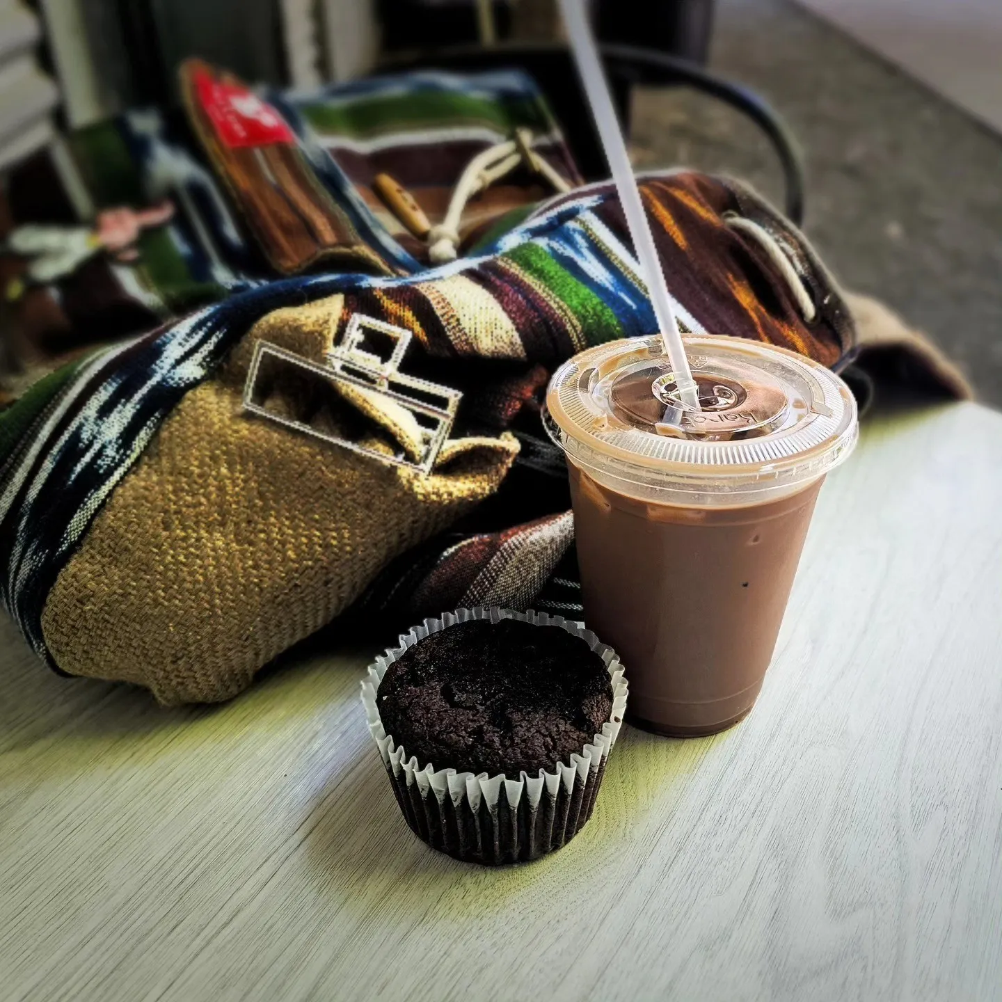 An iced drink and a chocolate muffin sit beside a patterned bag on a table.