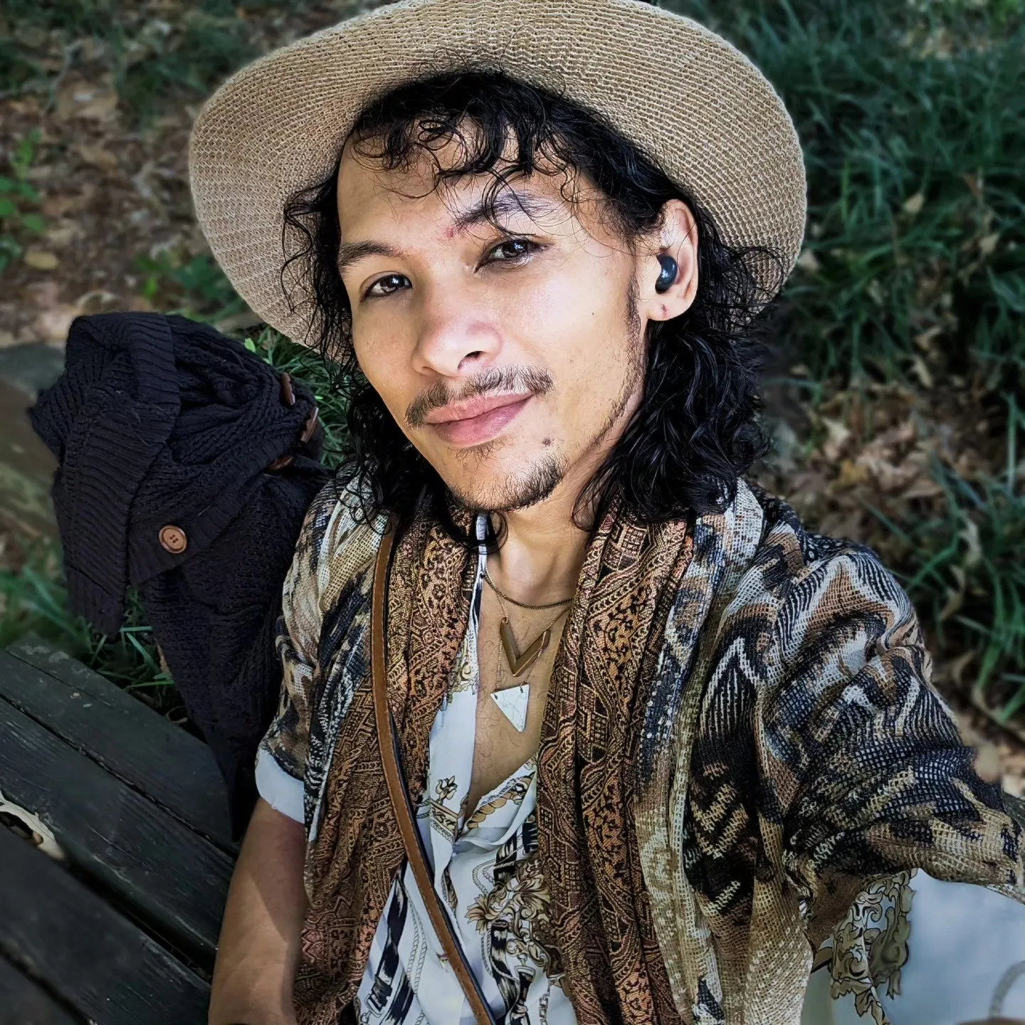 A person in a straw hat and patterned shirt takes a portrait beside a backpack outdoors.