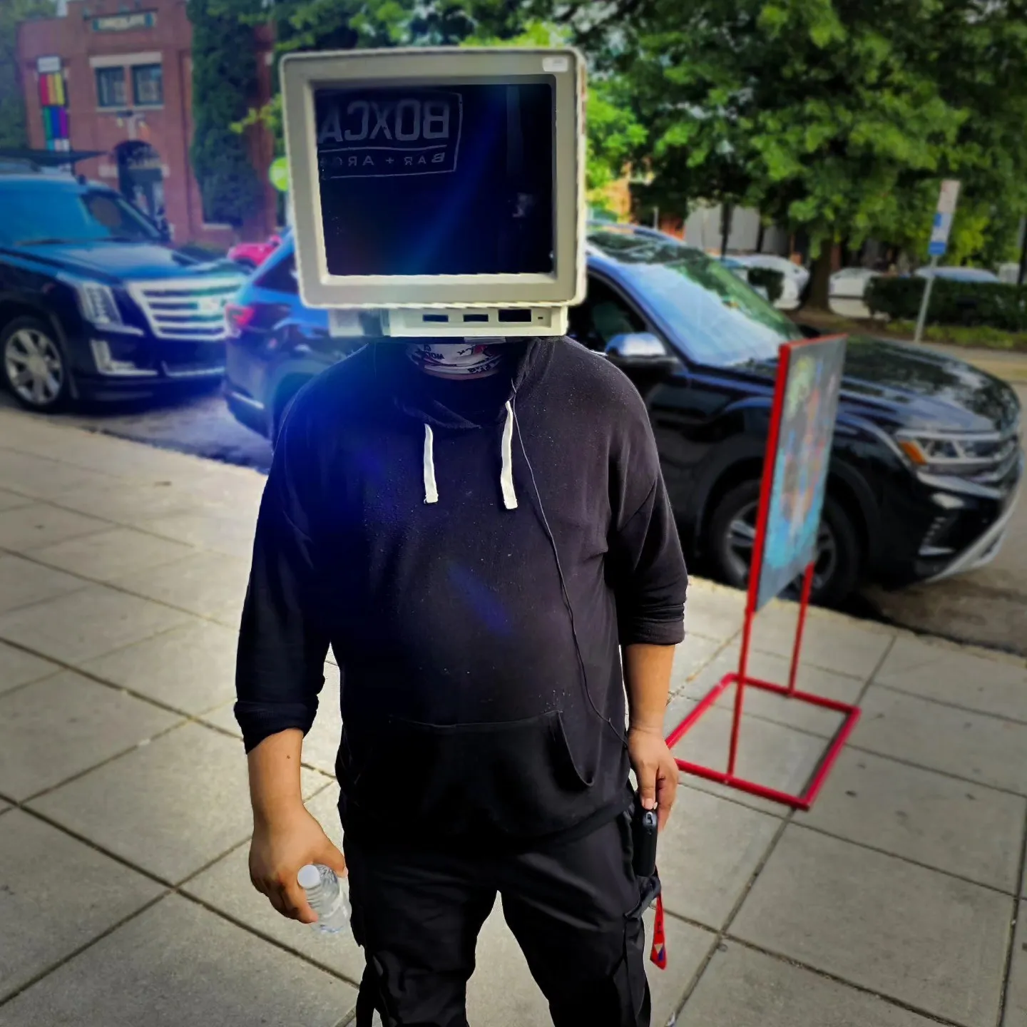 A person in dark clothes stands on a sidewalk wearing a computer monitor over their head.