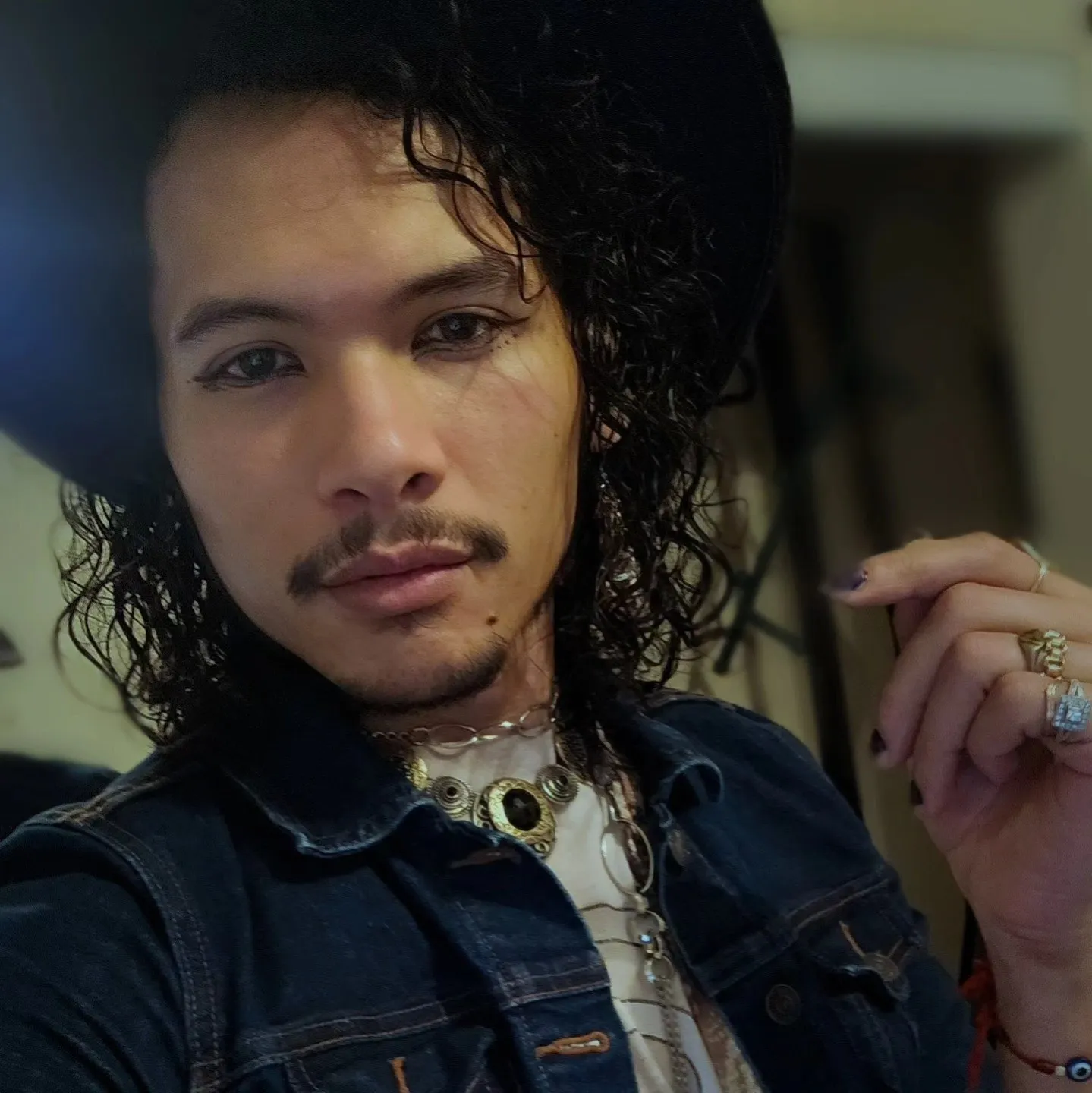 A close portrait of a person with dark curls, eyeliner and a decorative necklace.