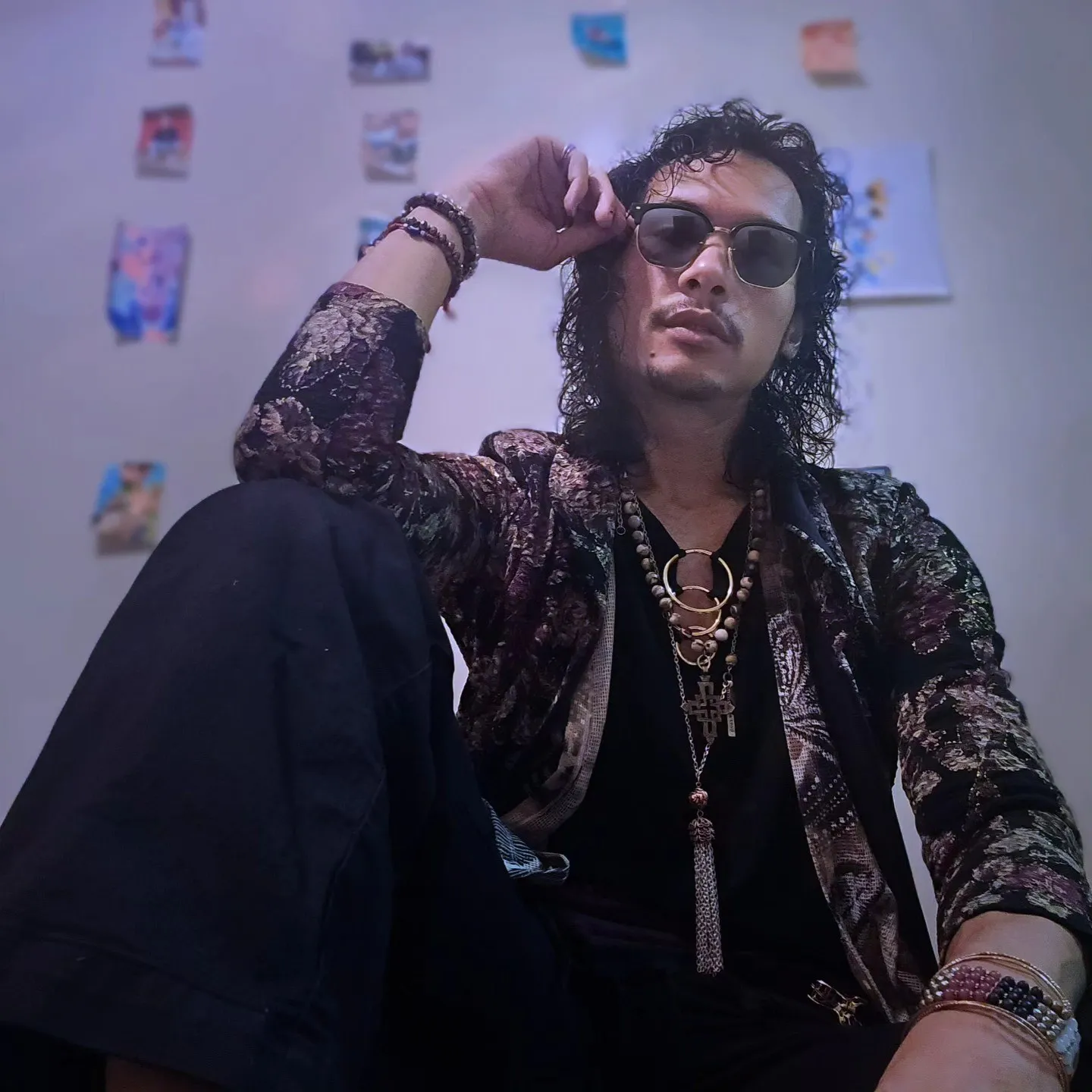 A seated person in sunglasses, a patterned jacket and layered necklaces raises a hand to their glasses.