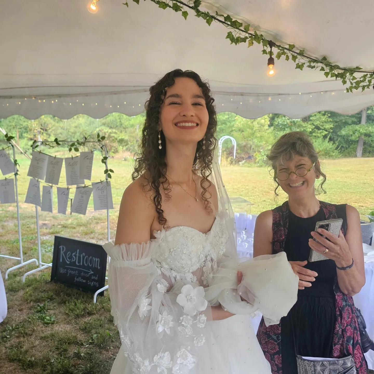 A smiling person in a white wedding dress stands beside a guest under a reception tent.
