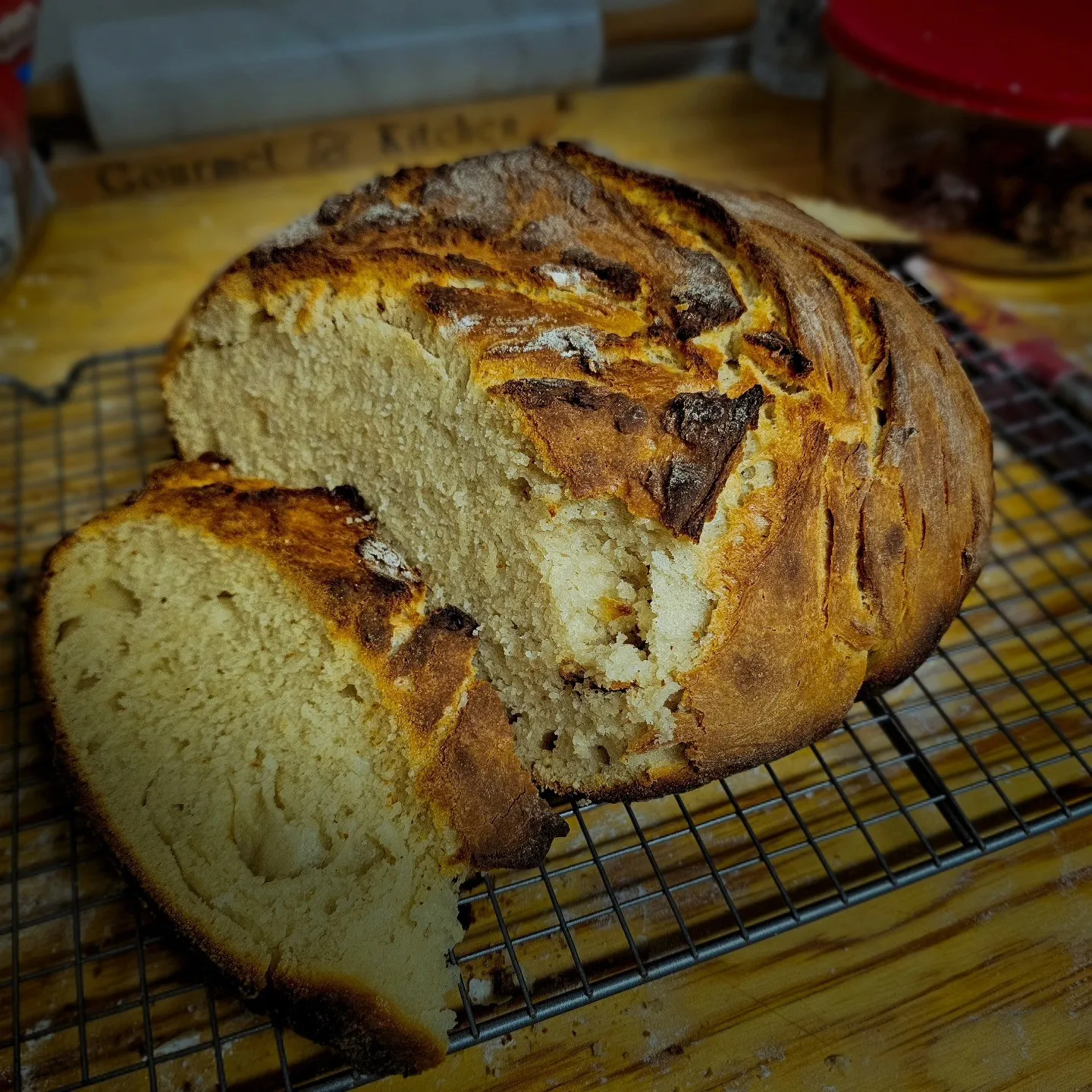 A round, deeply browned loaf of bread cools on a rack with one slice cut away.