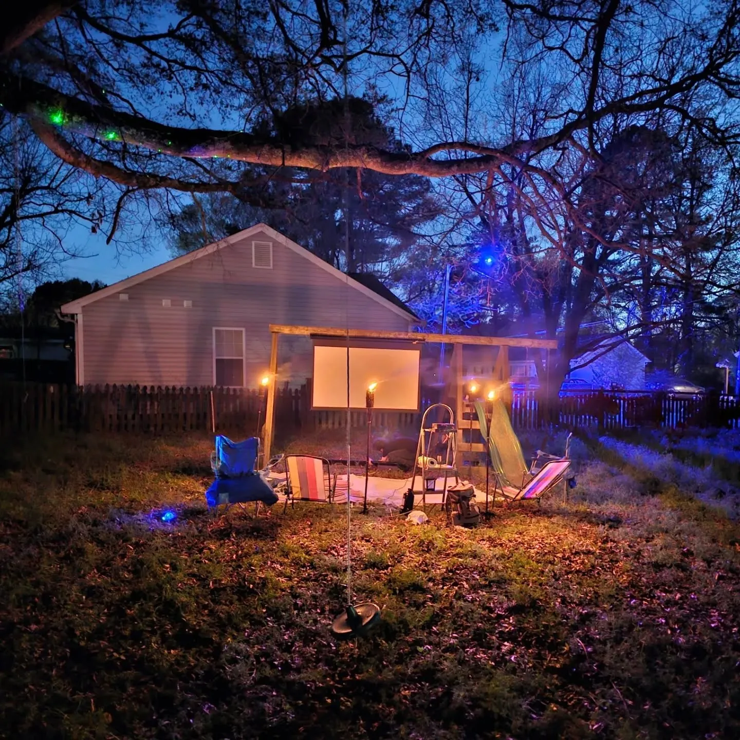 A backyard at dusk with colored lights strung through a tree and a small illuminated canopy.