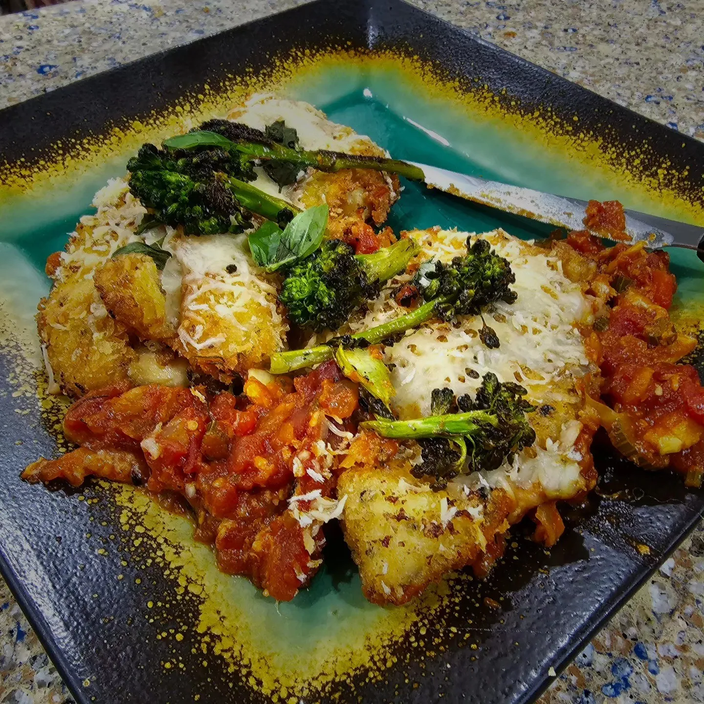 A baked dish topped with melted cheese and broccoli on a turquoise and black plate.