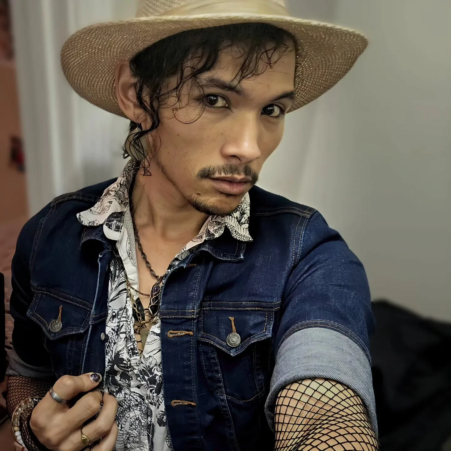 A person wearing a straw hat and denim jacket takes a portrait indoors.