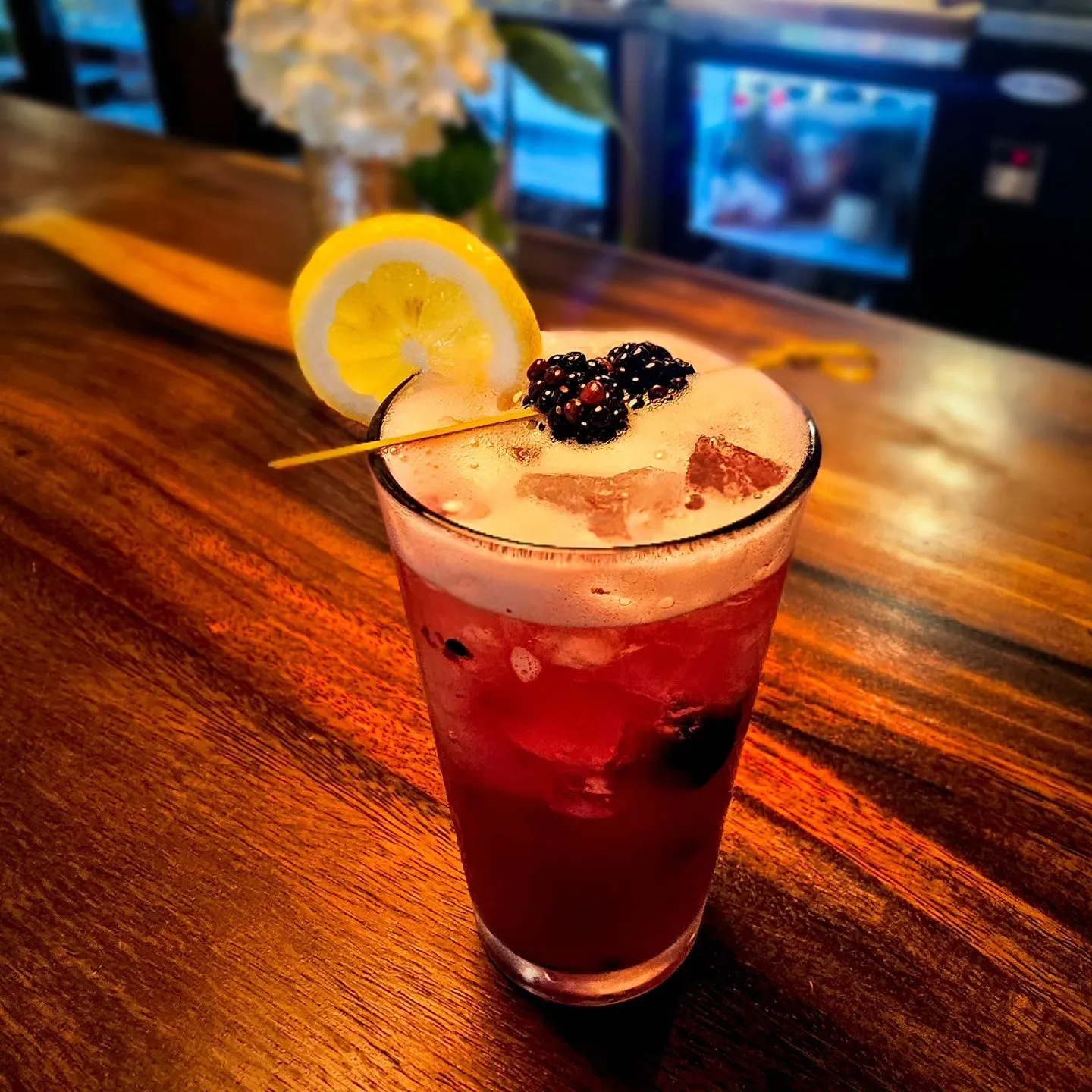 A tall red cocktail topped with foam, a lemon slice and a blackberry on a bar.