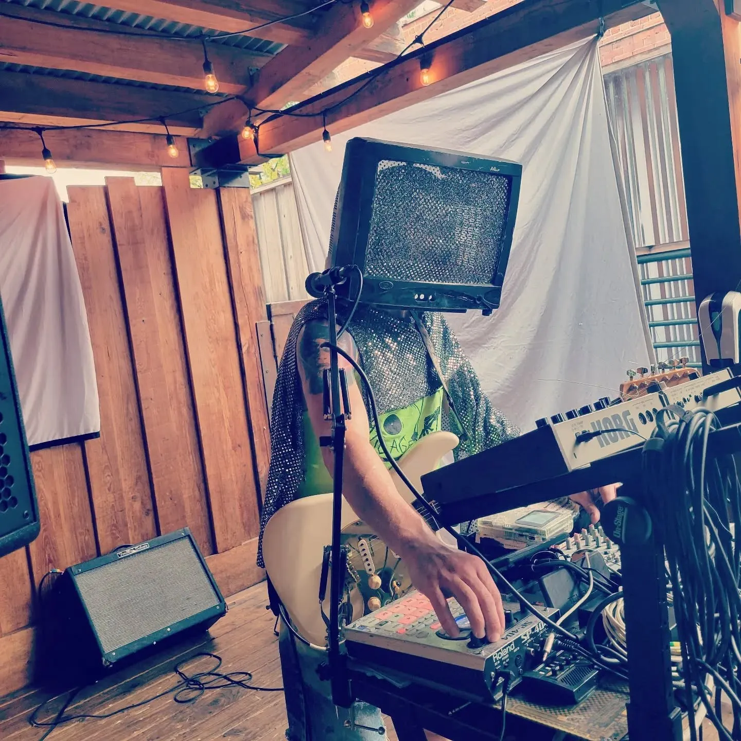 A performer wearing a monitor over their head adjusts electronic music equipment beside a keyboard.