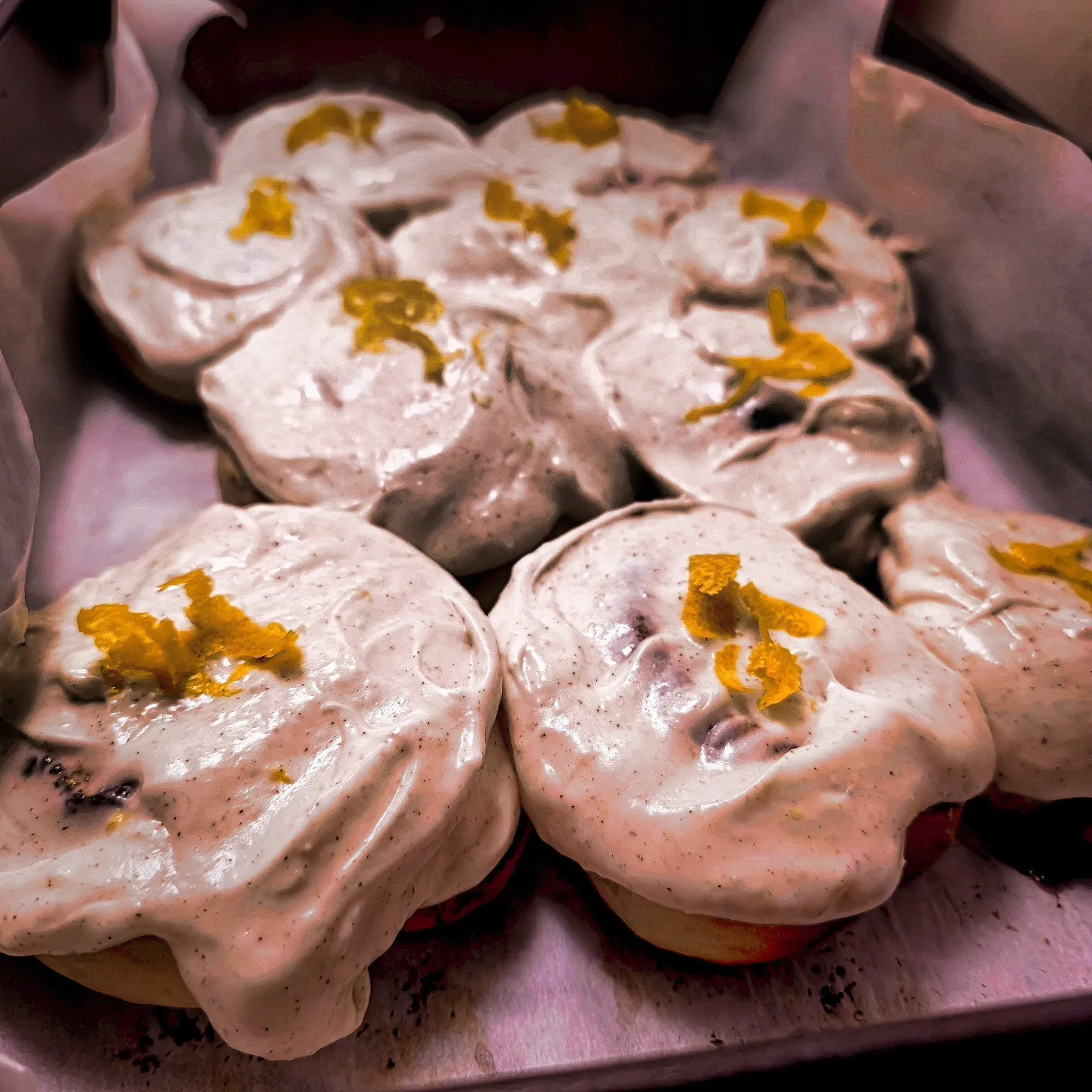 Frosted rolls topped with small curls of orange zest fill a baking tray.