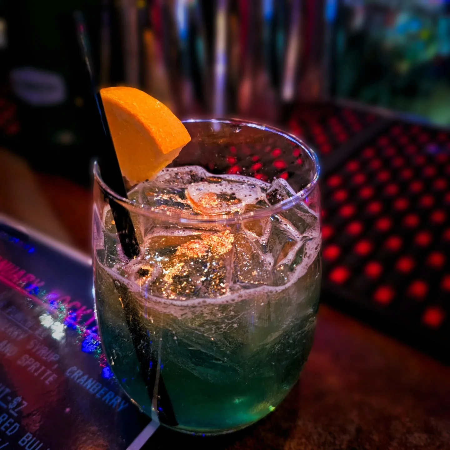 A blue-green iced drink with an orange wedge sits on a dimly lit bar.
