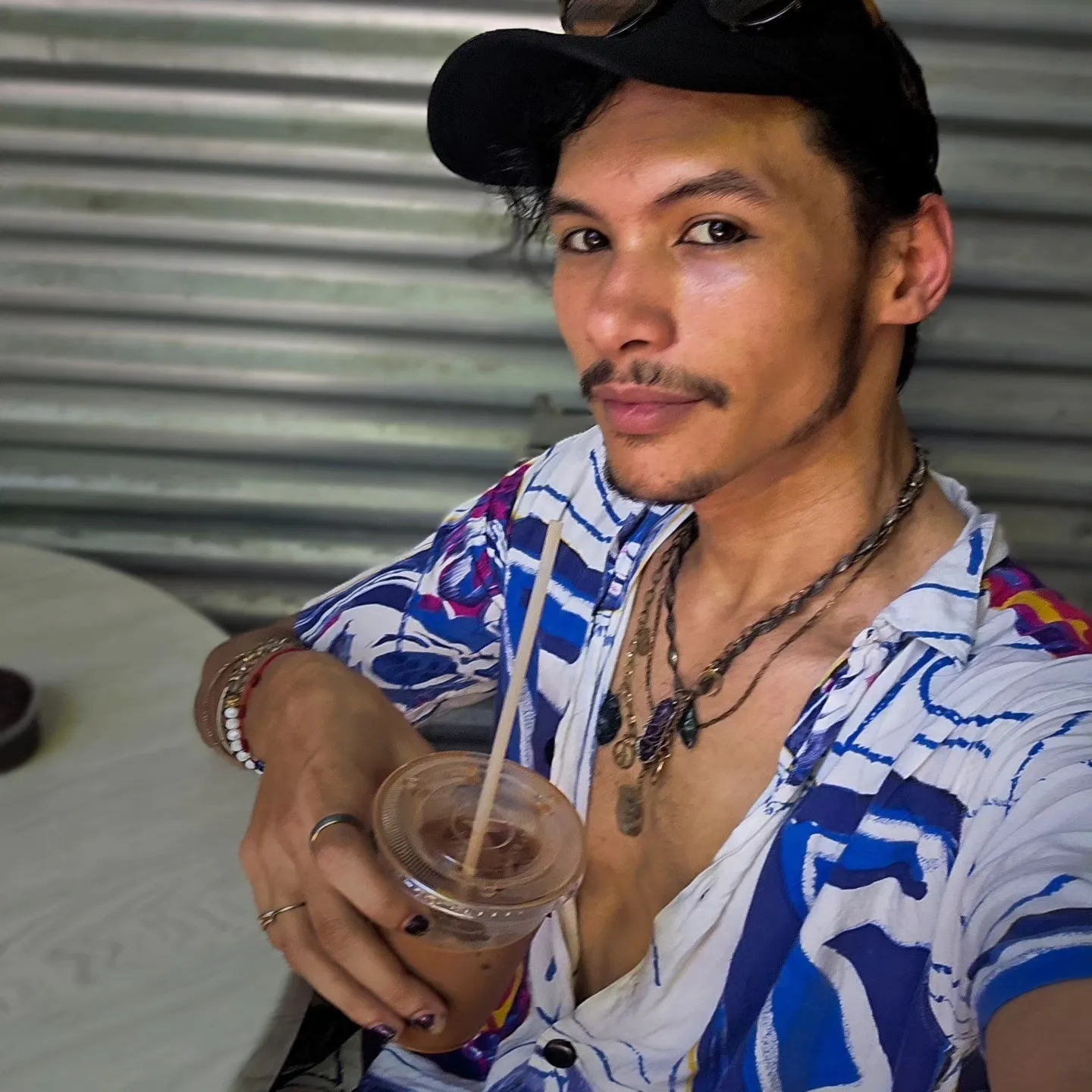 A person wearing a black cap and a blue-and-white patterned shirt holds an iced drink.
