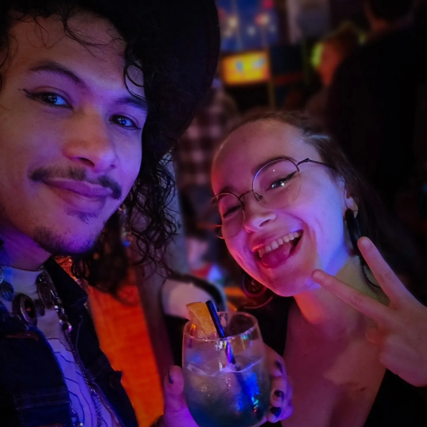Two people smile for a selfie in colorful bar lighting, one holding a drink.