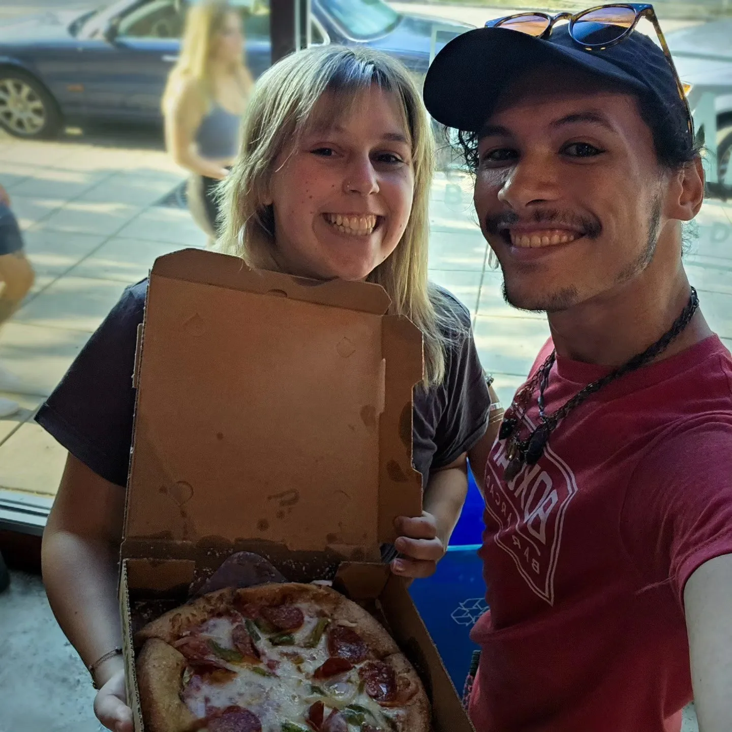 Two smiling people hold open a pizza box for a selfie.