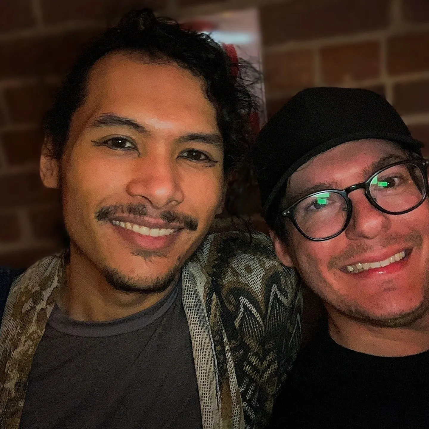 Two smiling people lean together for a close selfie against a brick wall.