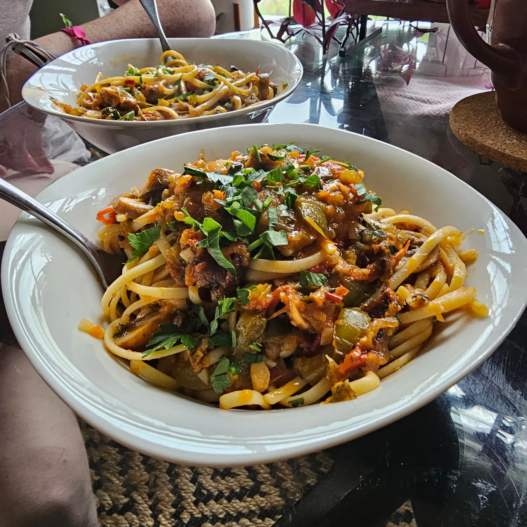Two bowls of pasta on a glass table, with potted plants, chairs, and the room around them still in view.
